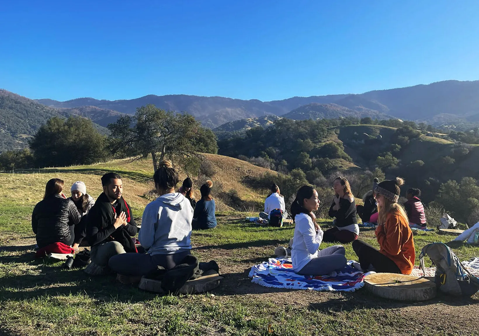People sitting on blankets on a grassy hilltop with mountains in the background.