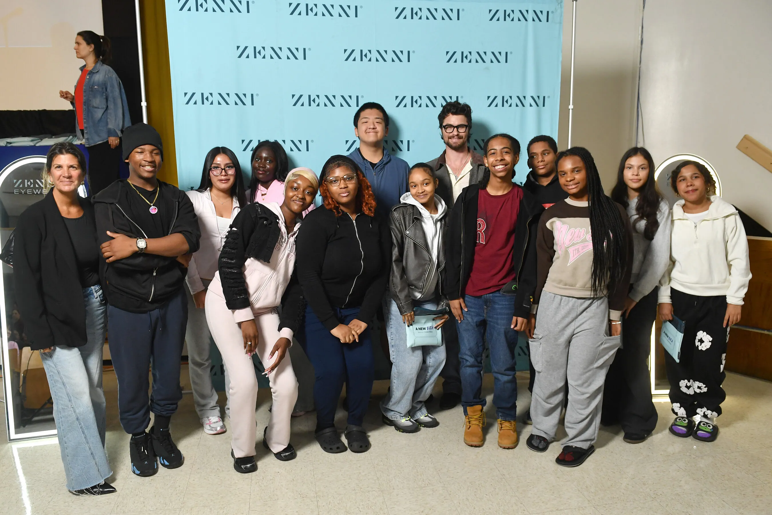 A group of adults and young people pose in front of a Zenni backdrop.
