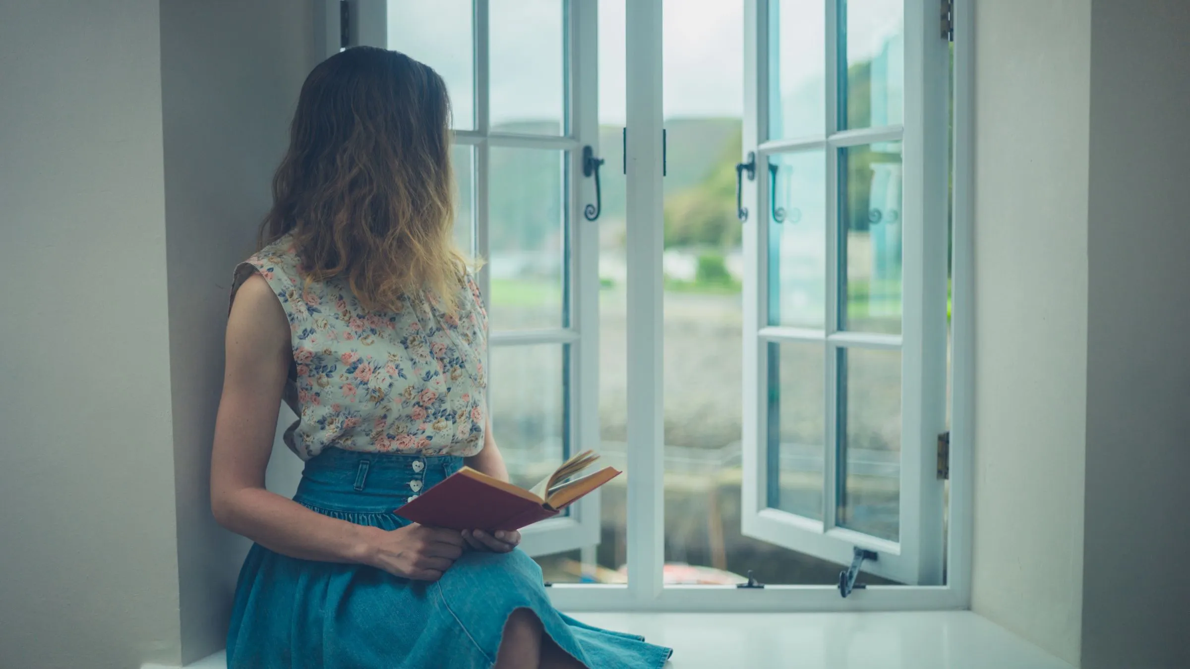 A woman holding a book beside an open window.