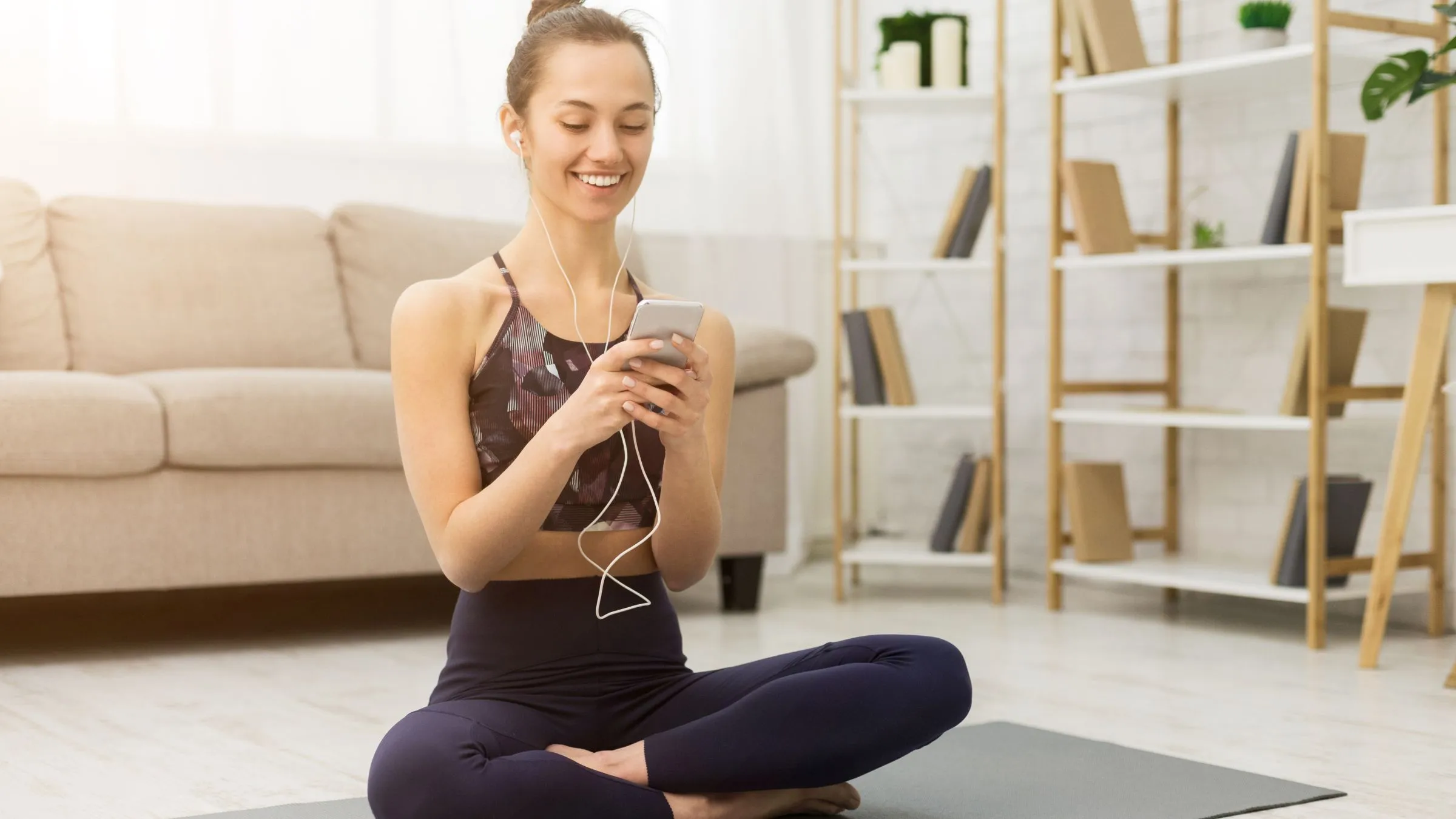 A smiling woman on a yoga mat at home looking at her phone with earphones.