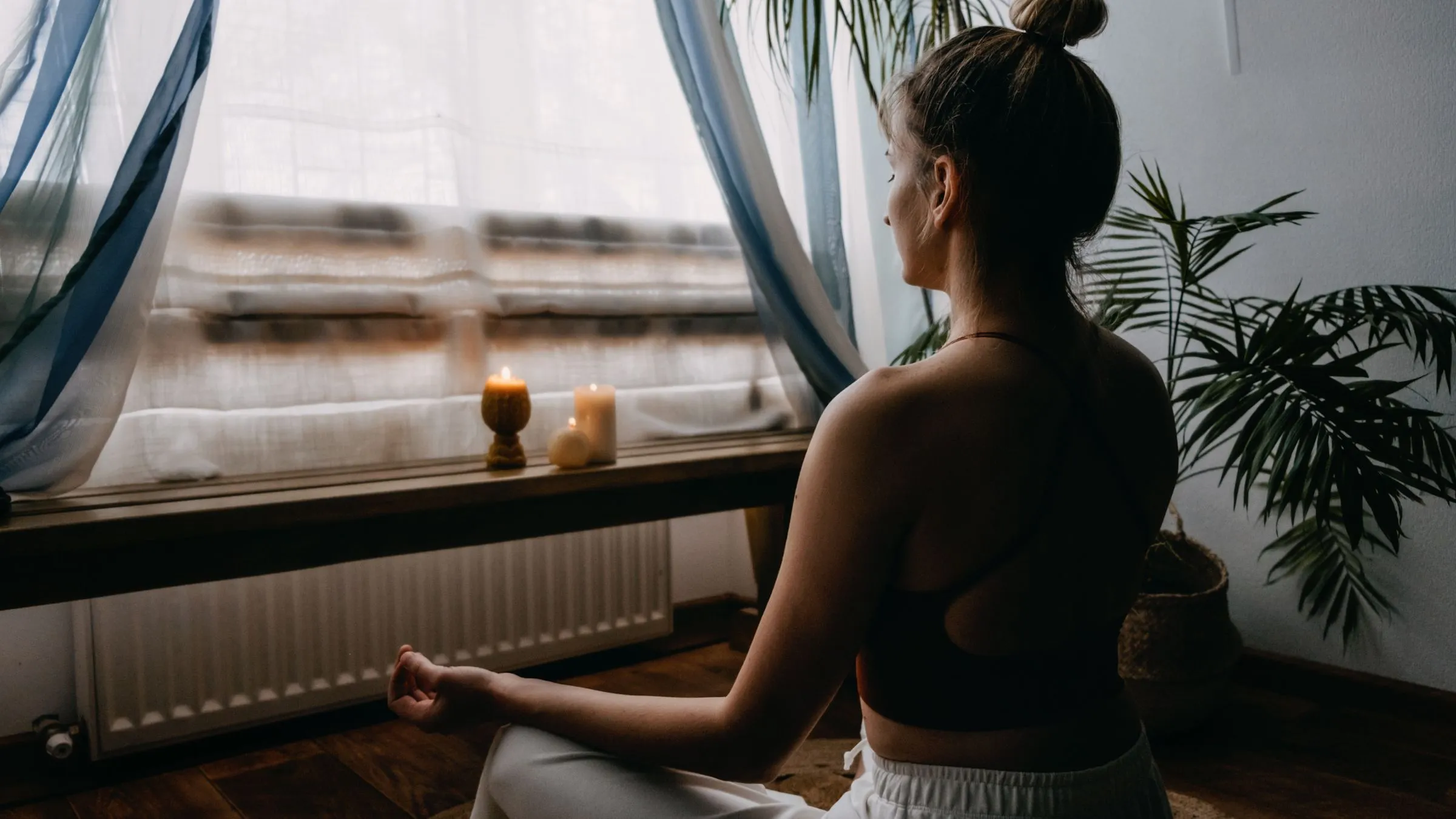A woman meditating cross-legged at home facing a window with candles.