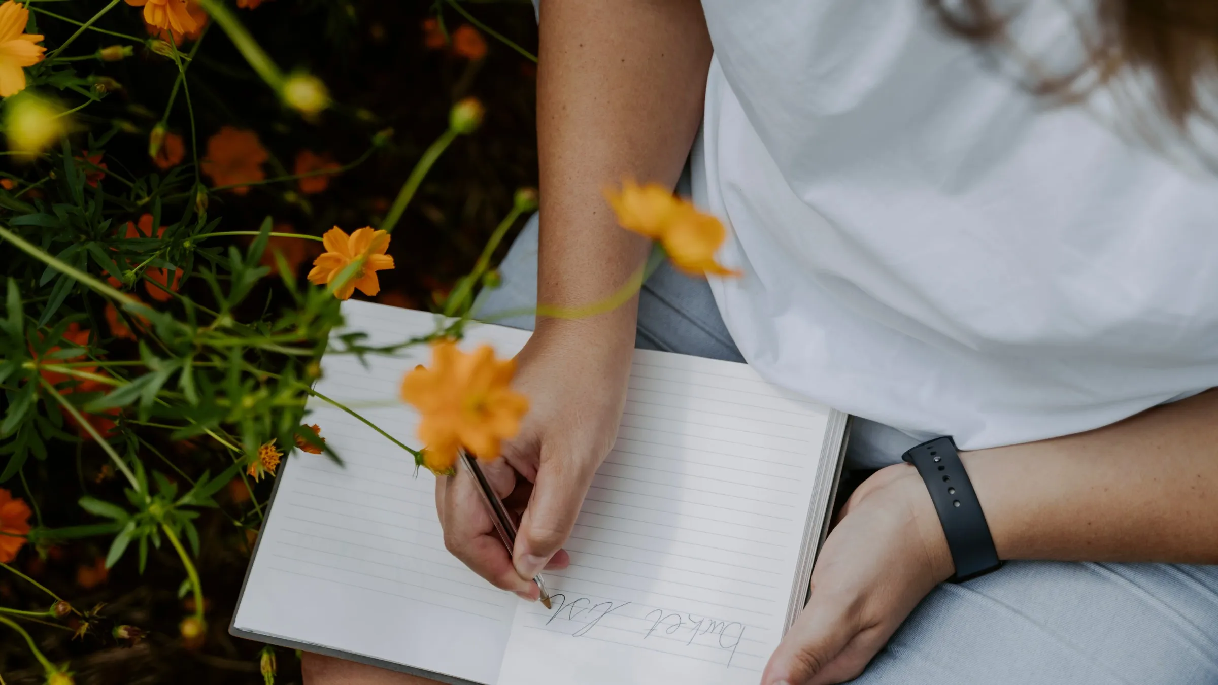 A woman in a white T-shirt writing "Bucket list" in an open notebook beside orange flowers.