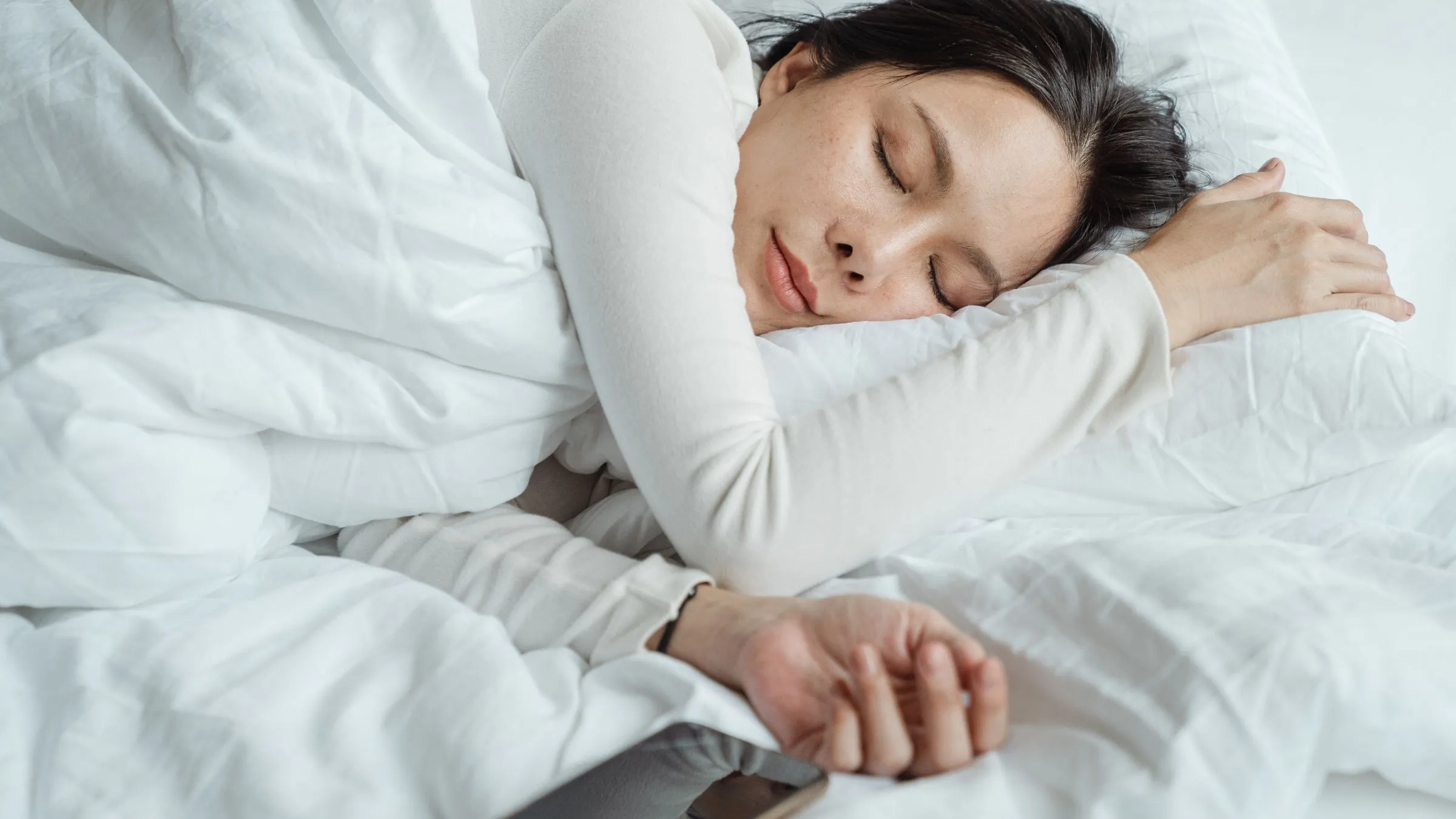 A woman asleep in white bedding with a phone beside her hand.